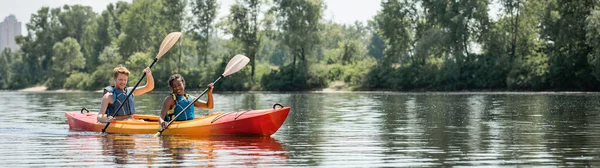 Alegre mujer afroamericana y joven hombre deportivo en chalecos salvavidas pasar tiempo en el pintoresco río mientras navega en kayak a lo largo de la orilla verde, bandera - foto de stock