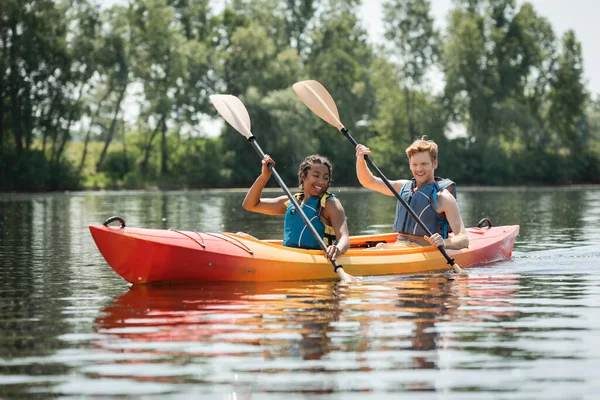 Affascinante donna africana americana con giovane e rossa uomo in giubbotti di salvataggio sorridente mentre pagaia in kayak sportivo sul lago con alberi verdi sulla riva in estate — Foto stock