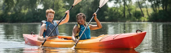 Amigos multiétnicos sonrientes y activos en chalecos salvavidas remando en kayak deportivo durante el fin de semana de recreación acuática en el pintoresco río en el día de verano, bandera - foto de stock