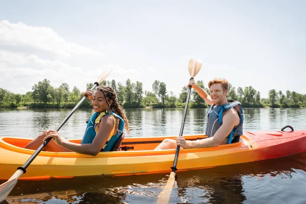 Feliz y encantadora mujer afroamericana con hombre joven y pelirrojo sosteniendo paletas y navegando en kayak deportivo en el lago escénico en fin de semana de verano activo - foto de stock