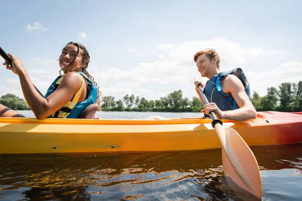 Sonriente y bonita mujer afroamericana navegando en kayak deportivo con el hombre pelirrojo activo en chaleco salvavidas durante las vacaciones de verano en el río con la orilla verde - foto de stock