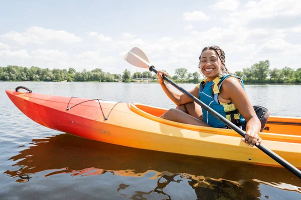 Насолоджена афроамериканська жінка в житті жилет веслування в спортивному каяку і посміхається на камеру на озері з зеленим мальовничим берегом на фоні влітку — стокове фото