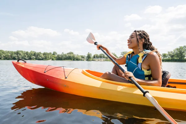 Вид збоку безтурботну і активну афроамериканську жінку в житті жилет тримає весло, плаваючи в каяку на озері з зеленим мальовничим берегом влітку — стокове фото
