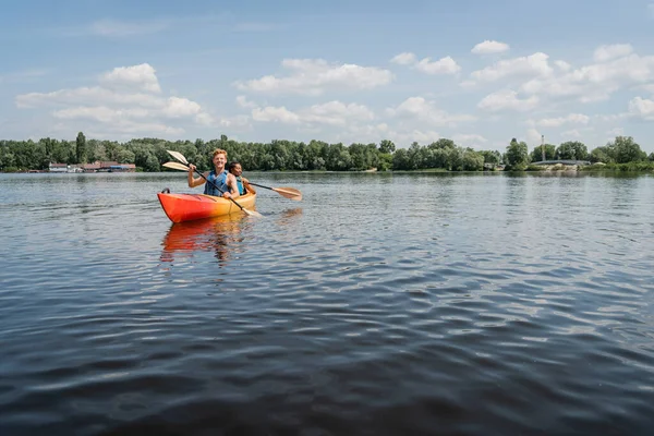Coppia multietnica attiva in giubbotti di sicurezza a vela in kayak sportivo sul pittoresco lago con riva verde sotto il cielo blu e nuvoloso durante la ricreazione dell'acqua nel fine settimana estivo — Foto stock