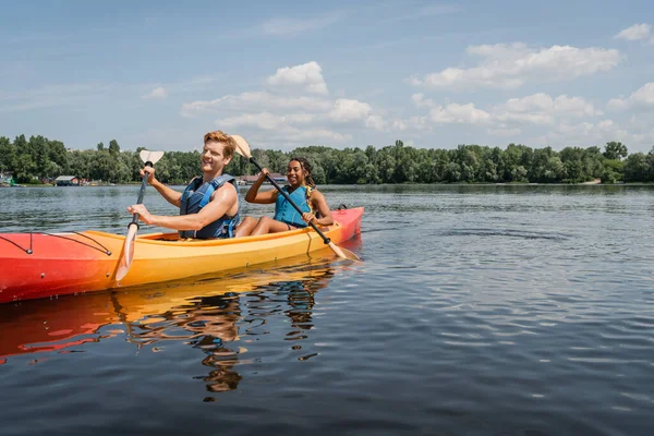 Happy and sportive interracial couple in life vests spending vacation on picturesque lake and sailing in kayak with paddles under blue sky with clouds — Stock Photo