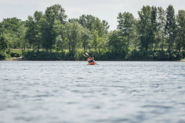 View from afar of active and interracial couple in life vests sailing in sportive kayak during recreation weekend on river with green bank during water recreation in summer — Stock Photo