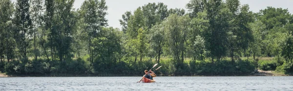 Vue de loin du couple actif interracial en gilets de sauvetage naviguant en kayak sportif avec pagaies près de la rivière avec des arbres verts le jour de l'été, bannière — Photo de stock