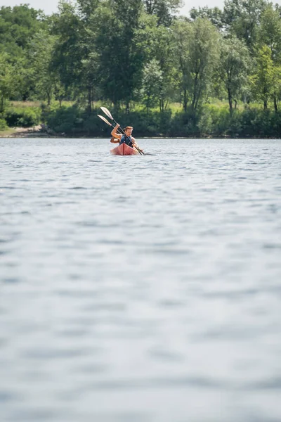 Blick aus der Ferne auf ein sportliches multiethnisches Paar in Schwimmwesten mit Paddeln beim Segeln im Kajak auf dem See mit grünem malerischem Ufer am Sommerwochenende — Stockfoto