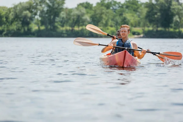 Young redhead man in life vest holding paddle and looking away while sailing in sportive kayak with african american woman during summer weekend on river — Stock Photo
