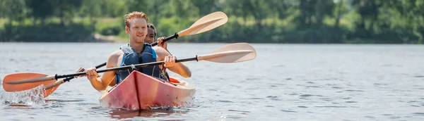 Молодий і спортивний рудий чоловік в житті жилет, дивлячись геть, тримаючи весло і плаваючи на байдарці з афроамериканкою на річці влітку, банер — стокове фото