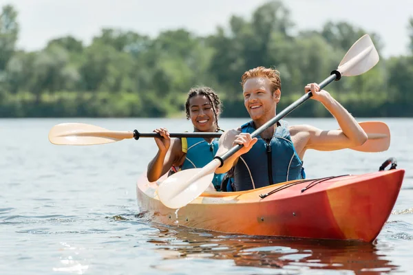 Hombre pelirrojo activo y encantadora mujer afroamericana en chalecos salvavidas pasar tiempo en el río mientras navega en kayak deportivo en el pintoresco lago sobre fondo borroso en verano - foto de stock