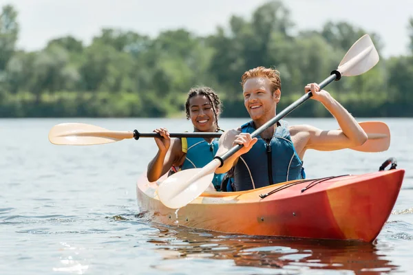 Active redhead man and charming african american woman in life vests spending time on river while sailing in sportive kayak on picturesque lake on blurred background in summer — Stock Photo