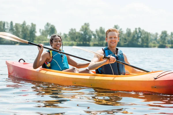 Young and carefree redhead man and pretty african american woman in life vests holding paddles and looking at camera while sailing in sportive kayak on river on summer day — Stock Photo