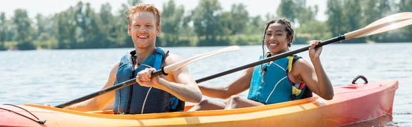Carefree interracial man and woman in life vests holding paddles and smiling at camera while sailing in sportive kayak on lake during summer weekend on blurred background, banner — Stock Photo