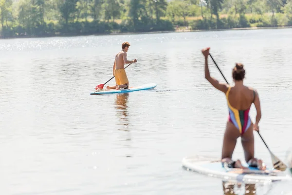 Jeune homme sportif naviguant sur une planche à soup sur un lac pittoresque près d'une femme afro-américaine en maillot de bain coloré pagayer au premier plan flou en été — Photo de stock