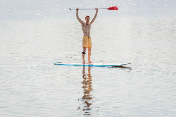 Volle Länge eines überglücklichen rothaarigen Mannes in gelben Badehosen mit Paddel in erhobenen Händen, während er am Sommerwochenende auf einem Surfbrett auf einem See mit ruhigem Wasser steht — Stockfoto