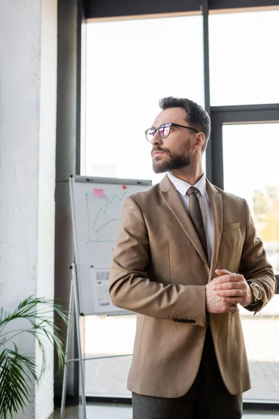 Вдумчивый бородатый корпоративный менеджер в модной формальной одежде и очках, стоящий рядом с размытым флип-чартом с бизнес-инфографикой и глядя в сторону в офисе — стоковое фото