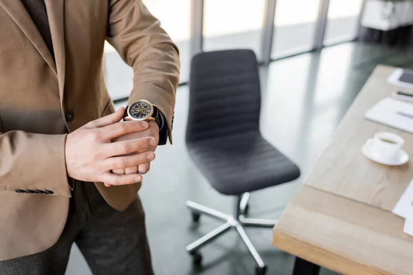 Vista parcial de empresario consumado en chaqueta beige y reloj de pulsera de lujo a la espera de cita cerca de la silla de oficina borrosa y taza de café en el escritorio - foto de stock