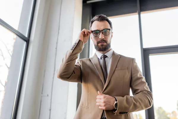 Vista basso angolo di ambizioso e di successo uomo d'affari barbuto in blazer beige elegante e cravatta occhiali di regolazione e guardando la fotocamera in ufficio moderno — Foto stock