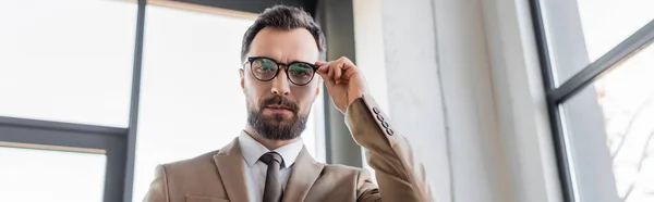 Entrepreneur beau et réussi en blazer élégant beige et cravate touchant lunettes élégantes et regardant la caméra dans le bureau contemporain, bannière — Photo de stock