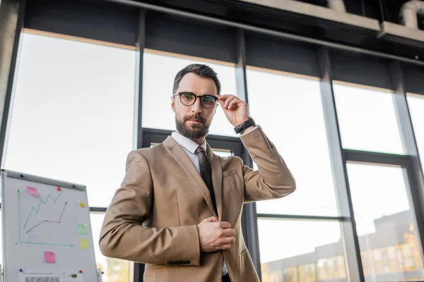 Confident manager in stylish formal wear adjusting eyeglasses and looking at camera near flip chart with business planning and analytics on background — Stock Photo