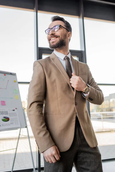 Low angle view of successful and happy corporate manager in beige blazer, tie and trendy eyeglasses looking away near flip chart with graphs and business strategy in office — Stock Photo