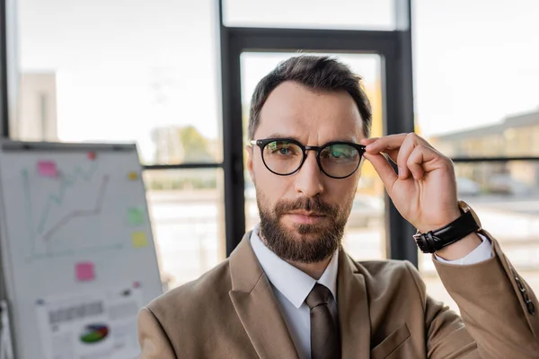 Бородатый успешный бизнесмен в бежевом пиджаке и галстуке, регулирующий стильные очки и смотрящий на камеру возле флип-чарта с графиками на размытом фоне — стоковое фото