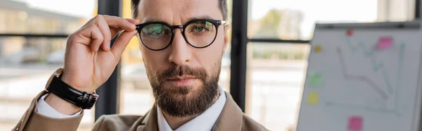 Retrato de gerente corporativo barbudo seguro y exitoso en desgaste formal y reloj de pulsera mirando a la cámara y ajustando gafas de moda en la oficina, pancarta - foto de stock