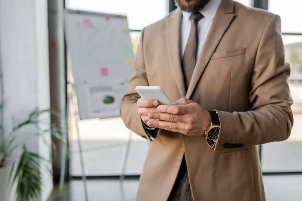 Partial view of elegant and accomplished businessman in beige blazer and tie using mobile phone near flip chart with strategy planning and graphs on blurred background — Stock Photo