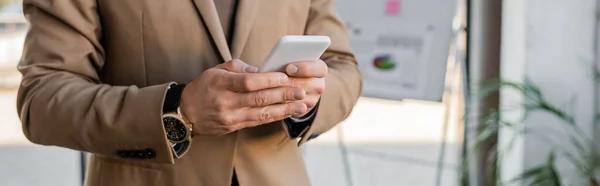 Cropped view of accomplished entrepreneur wearing beige blazer and stylish wristwatch, using mobile phone near flip chart with business analytics on blurred background, banner — Stock Photo