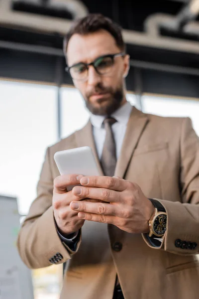 Low angle view of serious blurred businessman in luxury wristwatch, beige stylish blazer, eyeglasses and tie using mobile phone and thinking in office — Stock Photo