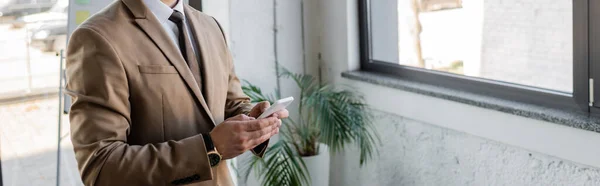 Vista parcial del gerente exitoso en blazer elegante beige y mensajería de corbata en el teléfono móvil en la oficina cerca de la planta en maceta sobre fondo borroso, bandera - foto de stock