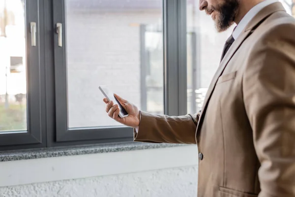Частичный вид бородатого бизнесмена в бежевом стильном блейзере, держащего мобильный телефон с пустым экраном, стоя рядом с большими окнами в современном офисе — стоковое фото