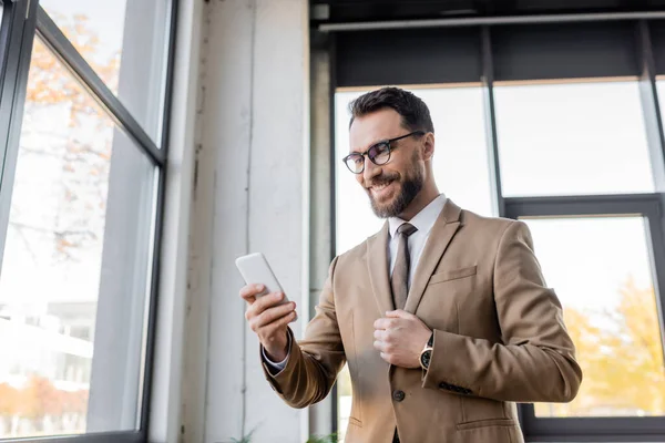 Charismatic and cheerful entrepreneur in trendy business attire such as beige blazer and eyeglasses standing near windows in office and looking at mobile phone — Stock Photo