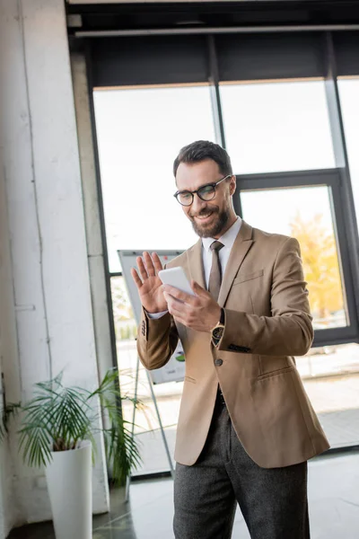 Gérant barbu joyeux en tenue de travail élégant et lunettes agitant la main lors d'un appel vidéo sur téléphone portable près de flip chart et plante en pot dans le bureau moderne — Photo de stock