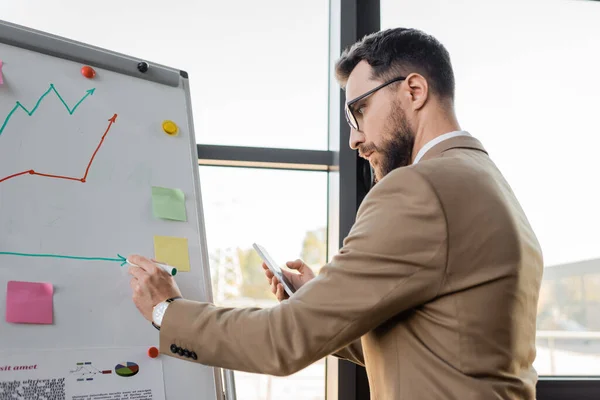 Side view of accomplished businessman in beige suit and eyeglasses holding mobile phone and drawing on flip chart while making productivity analysis in office — Stock Photo