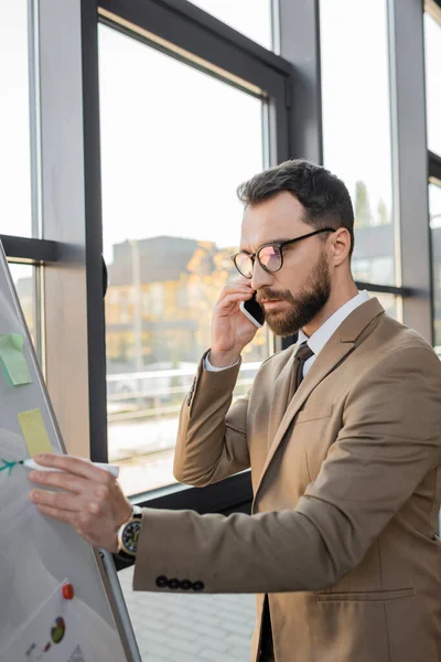 Serious bearded entrepreneur in stylish eyeglasses and beige blazer talking on mobile phone and drawing with marker on flip chart while doing business analytics in office — Stock Photo