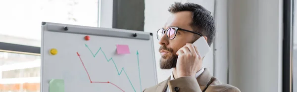 Portrait of thoughtful bearded businessman in stylish eyeglasses talking on mobile phone and looking away near flip chart with analytics in office, banner — Stock Photo