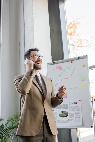 Joyful bearded manager in beige blazer, tie and eyeglasses standing with marker and talking on mobile phone near flip chart with business analytics and graphs in office — Stock Photo