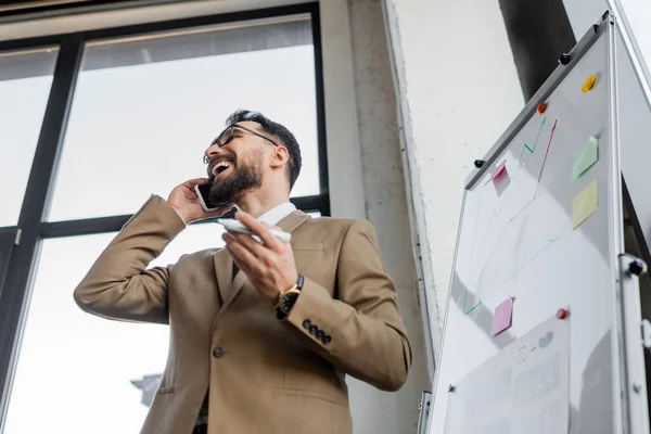 Low angle view of overjoyed bearded businessman in beige stylish blazer holding marker and talking on smartphone near flip chart with infographics and sticky notes in office — Stock Photo