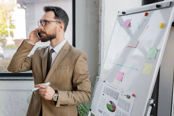 Pensativo hombre de negocios barbudo en blazer beige y gafas con marcador y mirando hacia otro lado mientras habla en el teléfono inteligente cerca de la investigación de mercado en el rotafolio en la oficina - foto de stock