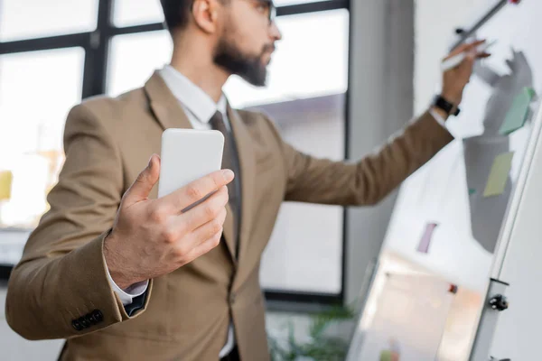 Homem de negócios em elegante blazer bege segurando smartphone e escrevendo em flip chart com marcador ao fazer a análise de produtividade no escritório, fundo borrado — Fotografia de Stock