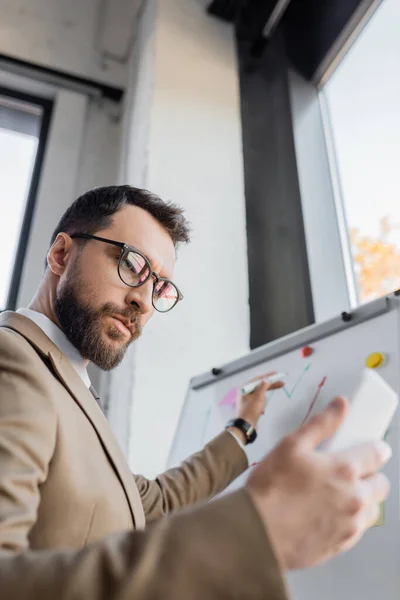 Low angle view of thoughtful and ambitious businessman in eyeglasses looking at mobile phone and holding marker near flip chart while doing market research in office — Stock Photo