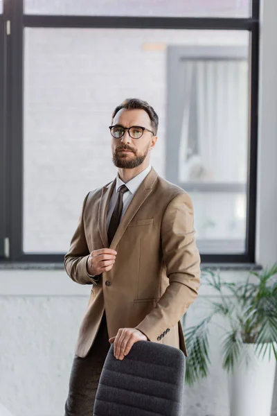 Уверенный и модный бизнесмен в очках, бежевый пиджак и галстук, стоящий возле стула и смотрящий в камеру в современном офисе на размытом фоне — стоковое фото