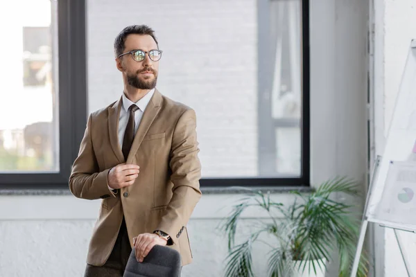 Уверенный и бородатый бизнесмен в бежевом пиджаке, галстуке и очках, стоящих возле офисного кресла, размытый флип-чарт и горшок с растением и отворачивающийся взгляд — стоковое фото
