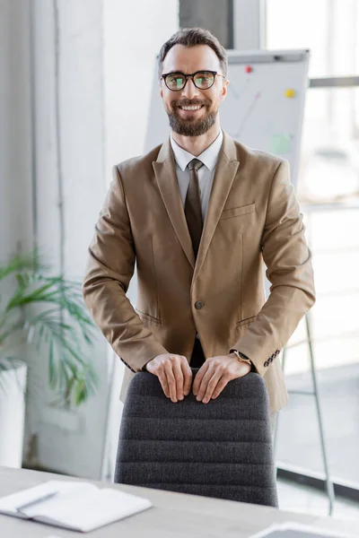 Alegre hombre de negocios guapo en blazer beige, corbata y gafas de vista de pie cerca de la silla de la oficina y sonriendo a la cámara delante del rotafolio sobre fondo borroso - foto de stock