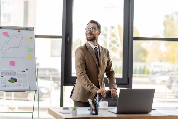Successful and charismatic businessman in stylish blazer and eyeglasses looking away near work desk with laptop, smartphone, coffee cup and stationery next to flip chart with infographics in office — Stock Photo