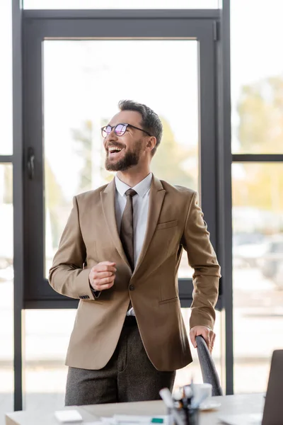 Homem de negócios satisfeito e muito feliz em blazer bege na moda, gravata e óculos olhando para longe perto de laptop borrado, smartphone e xícara de café na mesa de trabalho no escritório moderno — Fotografia de Stock