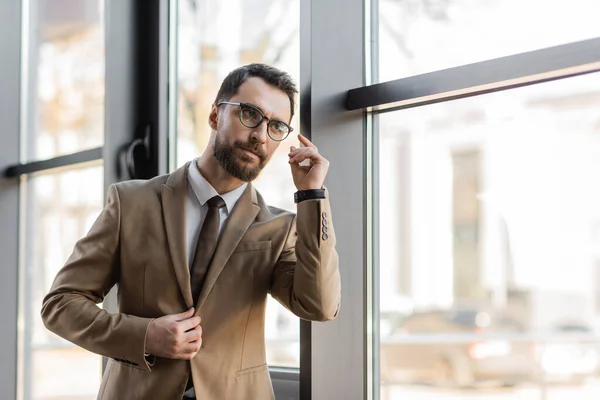 Emprendedor reflexivo y barbudo en traje de negocios de moda, tales como blazer beige, corbata y anteojos mirando lejos cerca de grandes ventanales en la oficina moderna - foto de stock