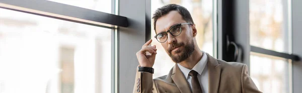 Homem de negócios barbudo em óculos da moda, blazer bege e gravata de mãos dadas perto do rosto, olhando para longe e pensando perto de janelas no escritório moderno, banner — Fotografia de Stock